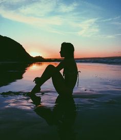 a woman sitting on the beach at sunset with her feet in the water looking out to sea