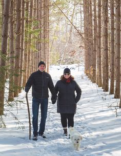 two people and a dog are walking through the snow in front of some tall trees