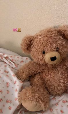 a brown teddy bear sitting on top of a bed next to a white wall with pink flowers