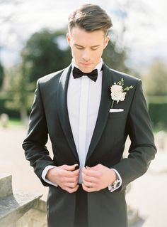 a man in a tuxedo is looking down at his bow tie while wearing a boutonniere