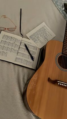 an acoustic guitar, notebooks and glasses are on a bed with sheets of music