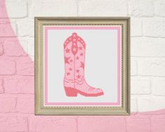 a cross stitch pattern of a pink cowboy boot on a white brick wall in front of a beige frame