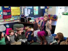 a woman standing in front of a classroom full of children sitting on the floor and talking to each other