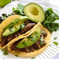 two shredded beef tacos with avocado and cilantro on a paper plate