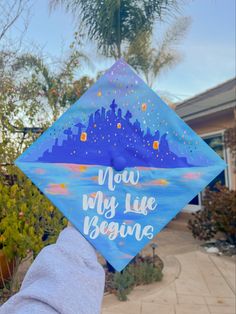 a blue graduation cap with the words now my life begins written on it in front of a palm tree