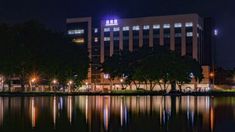 the building is lit up at night with lights reflecting in the water and trees around it