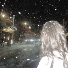 a woman standing in the middle of a snowy street at night with snow falling all around her