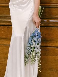 a woman in a white dress holding a blue and white flower bouquet next to a wooden wall