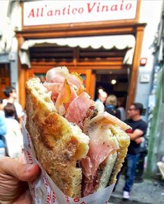 a person holding up a half eaten sandwich in front of a building with people walking by