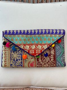 Embrace the beauty of artisan craftsmanship and make a meaningful impact with this stunning handcrafted clutch. Created by underprivileged women in India, this vibrant and intricately embroidered bag is not just a stylish accessory, but a symbol of empowerment and community support. Each clutch is a work of art, featuring a patchwork of rich, colorful fabrics adorned with intricate gold embroidery and playful pom poms that add a touch of whimsy. The envelope design is both elegant and practical, offering ample space for your essentials while ensuring you stand out in any crowd. By purchasing this clutch, you're not only adding a unique piece to your collection but also contributing to the livelihoods of talented artisans who pour their heart and soul into every stitch. These women, many fr Multicolor Embroidered Clutch For Festive Occasions, Traditional Multicolor Embroidered Clutch, Festive Multicolor Embroidered Clutch, Red Handmade Clutch For Festive Occasions, Handmade Red Clutch For Festive Occasions, Festive Handmade Red Clutch, Handmade Envelope Clutch As Gift, Handmade Envelope Clutch For Gift, Festival Multicolor Embroidery Handwork Clutch