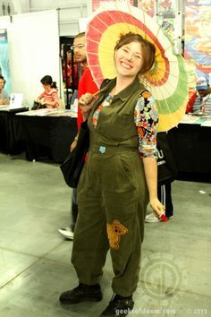 a woman in overalls holding an umbrella and smiling at the camera while standing next to a table