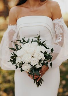 a woman in a white wedding dress holding a bridal bouquet with greenery on it