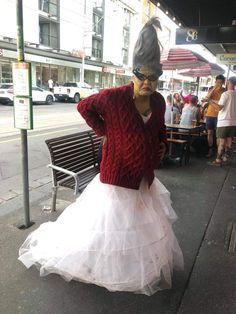 a woman in a white dress and red cardigan is standing on the sidewalk next to a bench