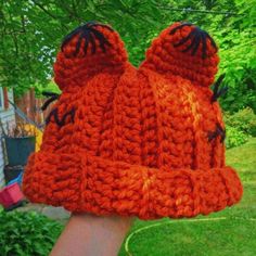 an orange crocheted hat is held up by someone's hand in the yard