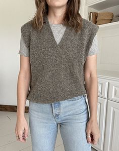 a woman standing in a kitchen wearing a sweater vest and jeans with her hands in her pockets