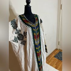 Ethiopian And Eritrean Traditional Dress (Habesh Kemis Gonder Kemis)Garment Featuring Colorful And Intricate Embroidery. This Style Is Often Seen In Ethiopian Or Eritrean Cultural Clothing, Known As "Habesha Kemis." The Dress Is Typically Worn By Women For Special Occasions And Celebrations. The Garment In Likely White Cotton Or Linen And Adorned With Detailed Patterns Along The Neckline, Cuffs, And Front Panel, Reflecting The Rich Textile Heritage Of The Region. Nepali Dhaka Dress, Fitted White Kaftan For Transitional Season, White Long Sleeve Transitional Kaftan, Fitted Multicolor Kaftan With Traditional Patterns, White Long Sleeve Dress With Traditional Patterns, Fitted Multicolor Kaftan For Traditional Ceremonies, Traditional White Thobe With Patterns, Traditional White Long Sleeve Habesha Kemis, Embroidered White Kaftan For Transitional Season