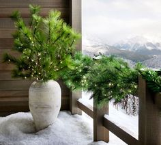 a white vase filled with pine branches sitting on top of a snow covered floor next to a window