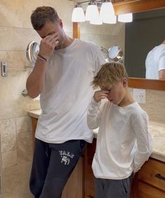 a man standing in front of a bathroom mirror next to a little boy who is covering his face