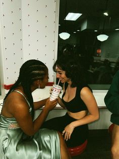 two women sitting next to each other in front of a window drinking from paper cups