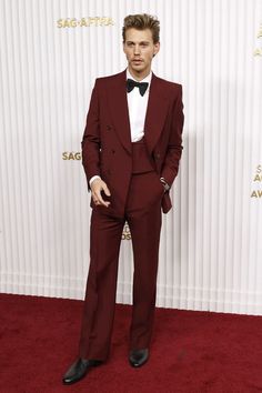 a man in a red suit and bow tie standing on a red carpet with his hands in his pockets