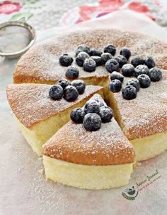 a cake with blueberries and powdered sugar sits on a tablecloth next to a spoon