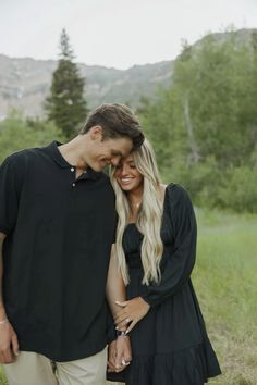 a man and woman standing next to each other in the grass with mountains in the background