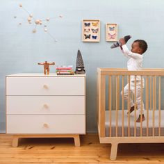 a baby standing on top of a crib next to a white dresser and chest