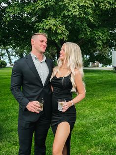 a man and woman standing next to each other in front of a tree holding wine glasses