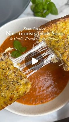 a person holding a piece of bread over a bowl of soup with tomato sauce and basil