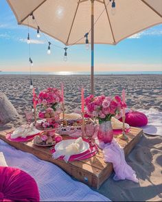 a table set up on the beach with pink flowers and desserts under an umbrella