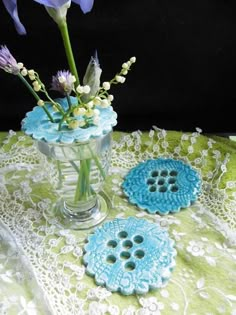 a vase filled with purple flowers sitting on top of a lace covered table cloth next to two blue buttons