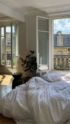 an unmade bed with white sheets and pillows in front of two large windows looking out onto the city