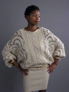 a woman standing in front of a gray background wearing a white sweater and beige skirt