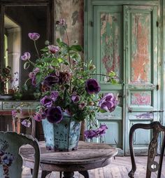 purple flowers are in a blue vase on an old table with two chairs around it