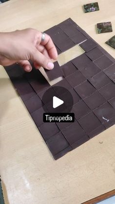 a person is making a mosaic with brown tiles on a wooden table and one hand is pointing at the tile