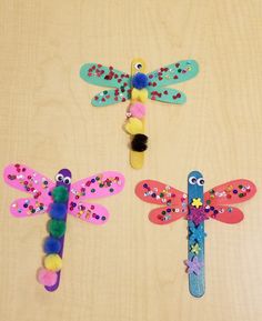 three dragonflys made out of colored paper on a wooden surface with beads and pom - poms