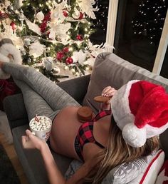 a woman laying on top of a couch wearing a santa hat and eating popcorn next to a christmas tree