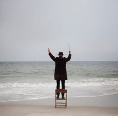 a man standing on top of a chair next to the ocean with his arms in the air