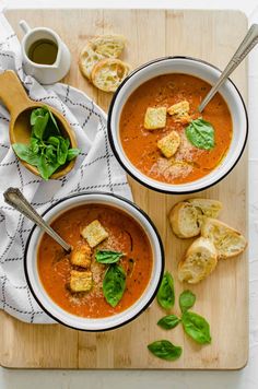 two bowls of tomato soup with bread and spinach on a cutting board next to a cup of coffee