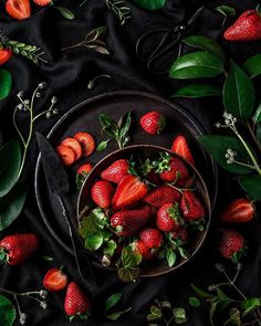 strawberries in a bowl surrounded by greenery and leaves on a black cloth background