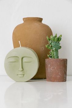 a clay face next to a potted plant and a ceramic candle holder on a white surface