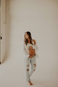 a pregnant woman standing in front of a white wall with her belly exposed and wearing ripped jeans