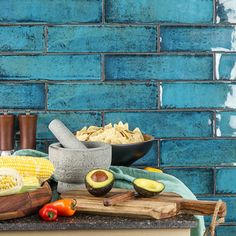 corn, avocado, and other foods are arranged on a wooden cutting board in front of a blue brick wall