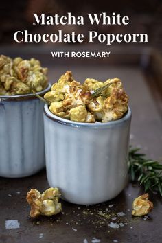 two silver cups filled with popcorn sitting on top of a counter next to rosemary sprigs