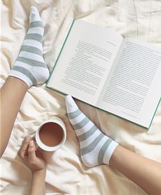 a woman laying in bed with her legs crossed and reading a book while holding a cup of coffee