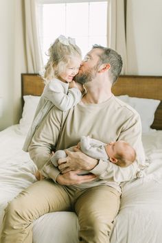 a man holding a baby while sitting on top of a bed next to a window