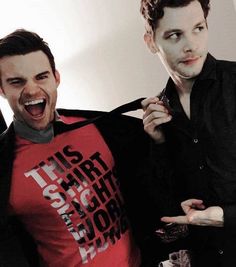 two men standing next to each other in front of a white wall and one is wearing a red shirt