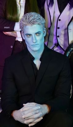 a man with white hair and blue skin sitting in front of other people wearing black suits