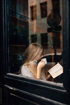 a woman sitting at a window reading a book and drinking from a cup by an open window