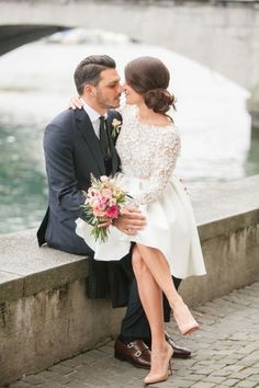 a bride and groom are sitting on a wall by the water